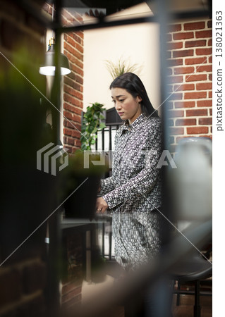 Female employee standing in brick wall workspace, organizing her desk with work belongings, preparing for the workday. Asian woman is on her feet by the table, arranging her office. Female employee standing in brick wall workspace, organizing her desk with work belongings, preparing for the workday. Asian woman is on her feet by the table, arranging her office. 138021363