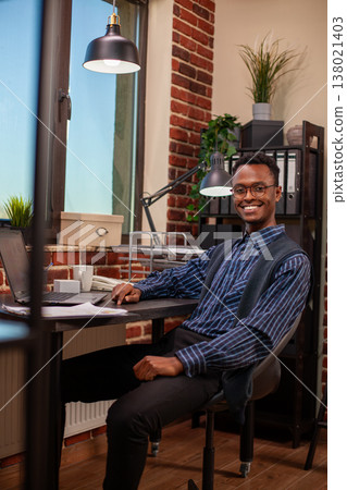 Portrait of african american man sitting at office desk and looking at camera. Black male entrepreneur with spectacles smiling brightly in home workspace with table arranged with laptop and documents. 138021403