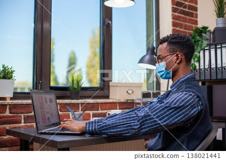 African american manager wearing earbuds, typing on digital device and researching marketing strategy at desk. Office worker with face mask, using laptop to review projects as he listens to podcasts. 138021441