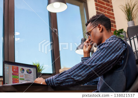 Male employee brainstorming at startup office desk, working on business project development with laptop. Black businessman touches his glasses and concentrates on screen displaying company analytics. Male employee brainstorming at startup office desk, working on business project development with laptop. Black businessman touches his glasses and concentrates on screen displaying company analytics. 138021537