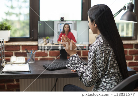 Rearview of young woman with an earbud, waving hello to coworker, commencing their online meeting on computer. Female entrepreneur making a greeting gesture to her business partner on a videocall. 138021577