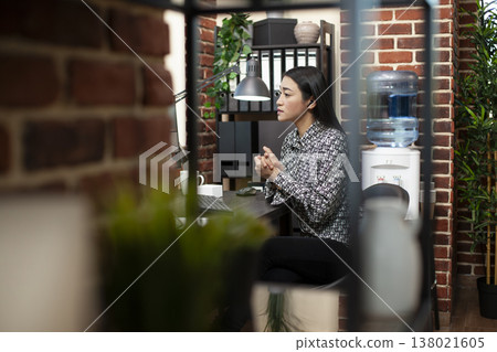 Asian female entrepreneur with earbuds, attends a virtual meeting from her office computer. Professional woman engages with colleagues during an online training session using her desktop pc. Asian female entrepreneur with earbuds, attends a virtual meeting from her office computer. Professional woman engages with colleagues during an online training session using her desktop pc. 138021605