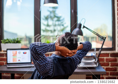 African american manager sits at desk, leaning back in his chair with hands behind head, enjoying quiet break in startup office. Black male employee looking out the window and reflecting on workday. 138021682