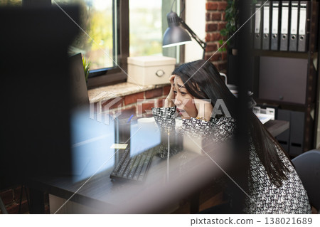 Asian female analyst appears stressed and overwhelmed in startup office. Young entrepreneur feeling tired, holding her head at desk and reviewing data on her computer in brick wall workplace. 138021689