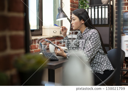 Female entrepreneur, concentrates on computer screen showing business analytics. Asian manager holding pen and clipboard with financial charts, comparing and analyzing information on desktop monitor. Female entrepreneur, concentrates on computer screen showing business analytics. Asian manager holding pen and clipboard with financial charts, comparing and analyzing information on desktop monitor. 138021709
