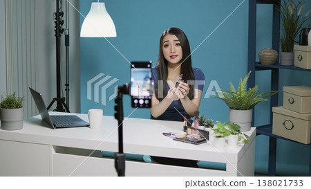 Content creator doing product review on camera in studio, filming video with makeup cosmetics. Female influencer recording lipstick recommendation vlog with smartphone for social media. 138021733