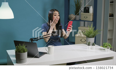 Female blogger answering social media comments on live podcast, talking to channel fans in studio. Influencer using smartphone technology and microphone to film online broadcast vlog. 138021734