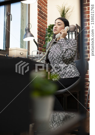 Female entrepreneur seated at office desk, sharing business project plans with client over the phone. Young asian manager holding landline receiver, calling her colleagues for a discussion. 138021768