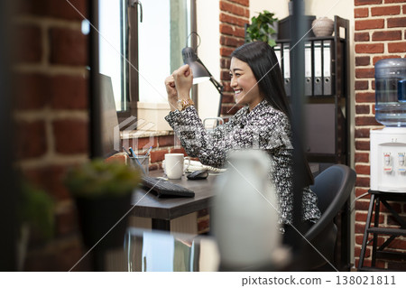 Asian female analyst triumphantly raises her fists in the office after achieving excellent outcomes for project presentation. Businesswoman smiling and celebrating a sealed business deal at her desk. Asian female analyst triumphantly raises her fists in the office after achieving excellent outcomes for project presentation. Businesswoman smiling and celebrating a sealed business deal at her desk. 138021811