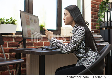 Female analyst pointing at computer screen, closely reviewing financial graphs in startup office. Young businesswoman concentrating on her desktop monitor, looking at company research information. 138021853