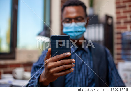 Businessman engaged in a videocall, holding his mobile device in brick wall office. Closeup of black male manager with face mask, grasping his smartphone and having virtual conversation with coworker. Businessman engaged in a videocall, holding his mobile device in brick wall office. Closeup of black male manager with face mask, grasping his smartphone and having virtual conversation with coworker. 138021856