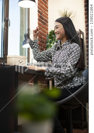 Asian woman celebrates at desk with her fist raised, after reading positive business outcomes on her computer. Excited female manager smiles, reflecting on success of company project. Asian woman celebrates at desk with her fist raised, after reading positive business outcomes on her computer. Excited female manager smiles, reflecting on success of company project. 138021864