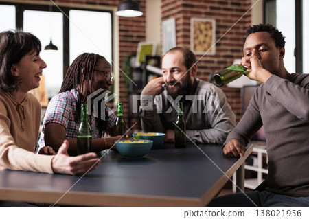 Happy multiracial friends sitting at home in living room while talking and enjoying snacks and beverages. Cheerful young adults having a conversation while relaxing with positive lifestyle. 138021956