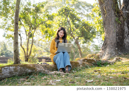 Pretty asian woman in yellow sweater looking at laptop while sitting on tree root with coffee cup. 138021975