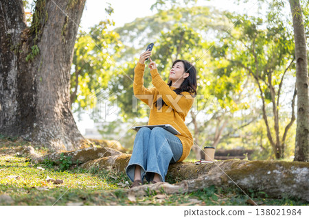 Pretty asian woman in yellow sweater holding phone taking picture while sitting on root under tree. Pretty asian woman in yellow sweater holding phone taking picture while sitting on root under tree. 138021984