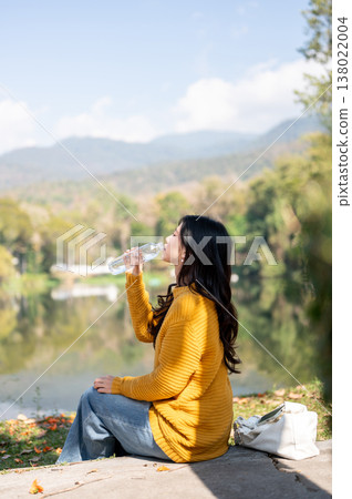 Side view of woman holding bottle drinking water while sitting with bag on sidewalk aside river lake Side view of woman holding bottle drinking water while sitting with bag on sidewalk aside river lake 138022004