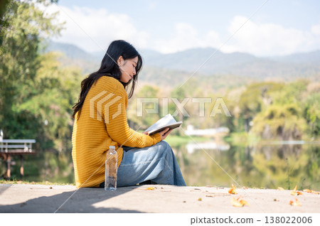 Asian woman reading a book while sits with water bottle on sidewalk aside river lake under sunlight. Asian woman reading a book while sits with water bottle on sidewalk aside river lake under sunlight. 138022006