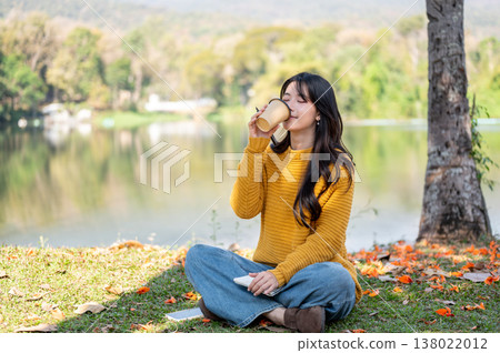 Pretty asian woman closing eyes drinking coffee sitting crossed legs on grass lawn aside river lake. Pretty asian woman closing eyes drinking coffee sitting crossed legs on grass lawn aside river lake. 138022012