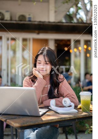 Happy asian woman resting head on hand looking at laptop aside green tea drink sitting at cafe table Happy asian woman resting head on hand looking at laptop aside green tea drink sitting at cafe table 138022025
