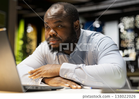African american worker in a dark office at night struggling to stay awake and finish his tasks, doing overtime alone at the workplace. Intense workload causes anxiety and work tension. 138022026