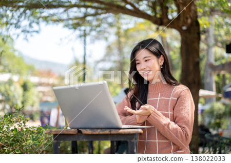 Happy asian woman talking explaining through a laptop while sitting at wooden table in cafe garden. 138022033