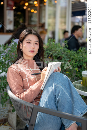 Pretty asian woman in pink sweater holding pen writing in notebook sits knees up on chair in garden 138022044