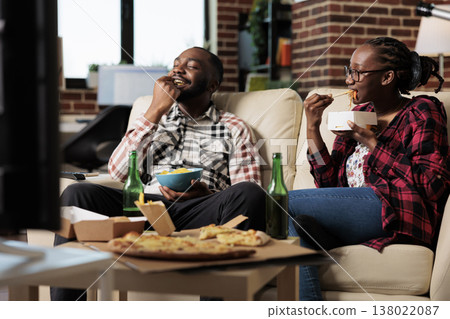 Young partners enjoying fast food delivery with movie on tv, having fun with leisure activity. Eating takeaway meal with noodles, pizza and chips, drinking beer from bottle. Watching television. Young partners enjoying fast food delivery with movie on tv, having fun with leisure activity. Eating takeaway meal with noodles, pizza and chips, drinking beer from bottle. Watching television. 138022087