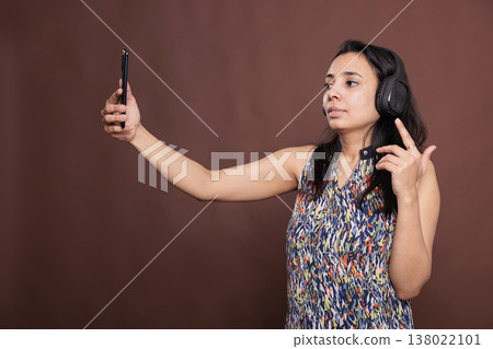Indian woman in headphones having videocall on smartphone, talking on teleconference. Person wearing wireless earphones, holding mobile phone, remote communication, online meeting Indian woman in headphones having videocall on smartphone, talking on teleconference. Person wearing wireless earphones, holding mobile phone, remote communication, online meeting 138022101