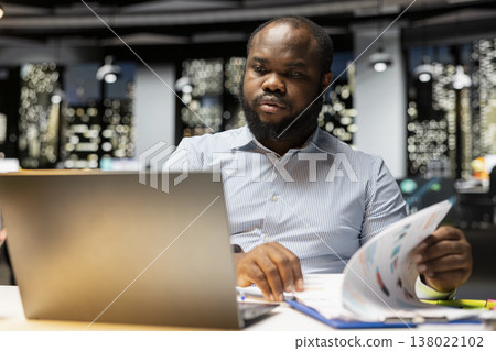 African american businessman verifying numbers and finance data in a dark office after hours, documenting updates and fixing the targets for upcoming business activity. Proofreading at night. African american businessman verifying numbers and finance data in a dark office after hours, documenting updates and fixing the targets for upcoming business activity. Proofreading at night. 138022102