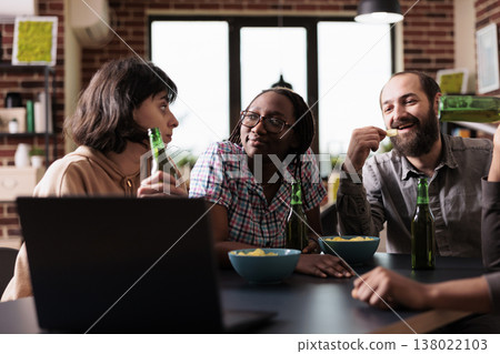 Positive diverse friends at home laughing and talking together while watching movies on laptop. Happy smiling people in living room enjoying fun leisure activity while having snacks. Positive diverse friends at home laughing and talking together while watching movies on laptop. Happy smiling people in living room enjoying fun leisure activity while having snacks. 138022103