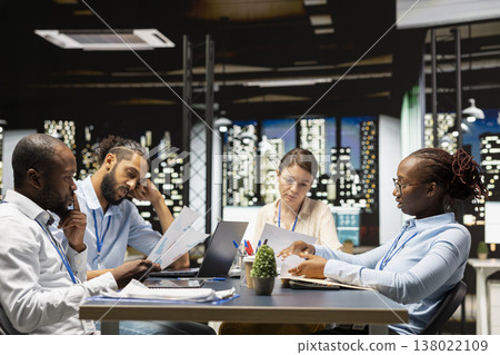 Diverse team of employees in office looking over paperwork documents late at night. Coworkers at work cooperating, analyzing company budget reports documentation files before deadline 138022109
