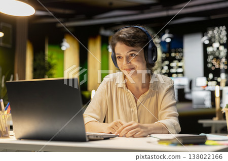 Businesswoman using headphones and working on tasks at desk, enjoys listening to focus music after hours in the office. Female adult creating an ambience and multitasking at night. 138022166