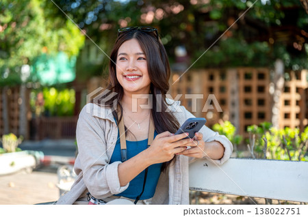 Smiling asian woman holding phone looking around while sitting or resting on sidewalk bench in park. 138022751