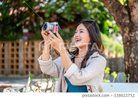 Smiling asian woman holding or raising camera taking picture while sitting on sidewalk bench in park Smiling asian woman holding or raising camera taking picture while sitting on sidewalk bench in park 138022752
