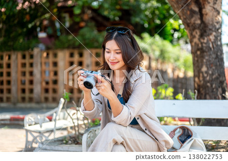 Pretty asian woman holding camera looking through photos while sitting crossed legs on bench in park Pretty asian woman holding camera looking through photos while sitting crossed legs on bench in park 138022753