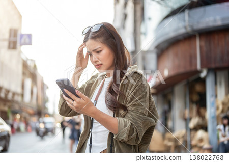 Confused asian woman scratching head looking at phone while standing by road street in local town. Confused asian woman scratching head looking at phone while standing by road street in local town. 138022768