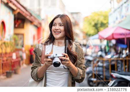 Pretty asian woman holding camera looking around standing or walking in local street market of town. Pretty asian woman holding camera looking around standing or walking in local street market of town. 138022776