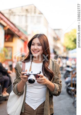 Pretty asian woman holding camera looking ahead standing or walking in local street market of town. Pretty asian woman holding camera looking ahead standing or walking in local street market of town. 138022777