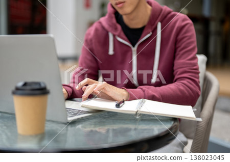 Close up of asian man student freelancer typing on laptop aside notebook and coffee at table in cafe Close up of asian man student freelancer typing on laptop aside notebook and coffee at table in cafe 138023045