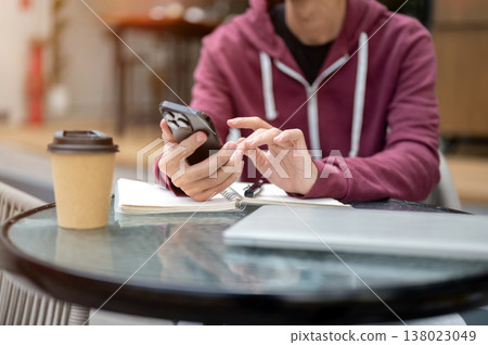 Close up of man student freelancer looking using phone over book aside laptop a coffee at cafe table 138023049