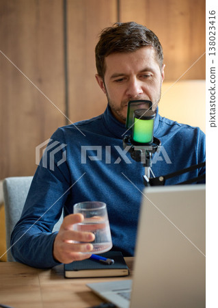 Man with glass of water speaking into microphone at laptop desk Man with glass of water speaking into microphone at laptop desk 138023416