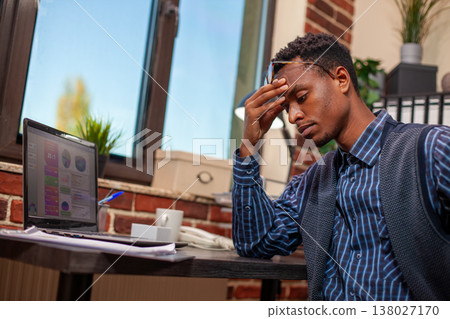 African American entrepreneur sits tiredly at desk, holding his head as he analyzes business project plans. Black male manager looking drained after demanding day at work in startup office. African American entrepreneur sits tiredly at desk, holding his head as he analyzes business project plans. Black male manager looking drained after demanding day at work in startup office. 138027170
