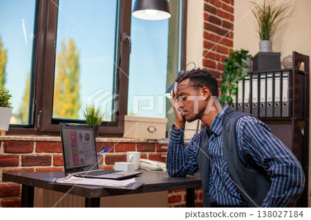 Stressed entrepreneur resting his face on his palm and appearing tired after long day at startup office. Businessman sitting at desk with laptop, rubs his eyes to be alert during work in workplace. 138027184