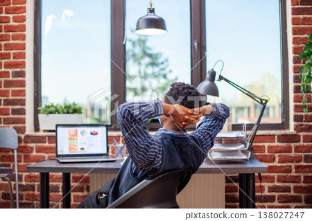 Black entrepreneur stretches his back in modern startup office, feeling accomplished after completing tasks. Male analyst with face mask, leaning in his chair and reflecting on productive day at work. Black entrepreneur stretches his back in modern startup office, feeling accomplished after completing tasks. Male analyst with face mask, leaning in his chair and reflecting on productive day at work. 138027247