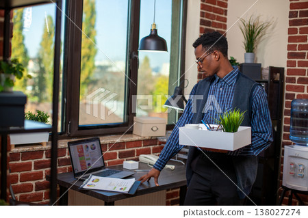 Male employee stands in workspace, holding box containing green plant and office materials, reviewing financial documents on desk. Black businessman checking company paperwork before leaving work. 138027274