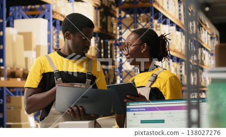 Warehouse staff overseeing parcels tracking info on laptop and tablet, preparing goods for outbound shipment. Organized depot with racks, cardboard boxes and processing tools. Camera B. 138027276