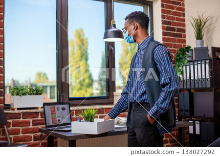 Business advisor wearing face mask stands at desk, arranging a box of work materials, including a plant pot. Black entrepreneur is reviewing important business project details and preparing to leave. Business advisor wearing face mask stands at desk, arranging a box of work materials, including a plant pot. Black entrepreneur is reviewing important business project details and preparing to leave. 138027292