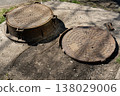 Rusty Open Manhole Cover and Raised Sewer Lid on Cracked Asphalt Sidewalk Surface 138029006