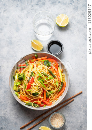 Stir fry wok noodles with vegetables in a plate on a light background with chopsticks, glass of water Stir fry wok noodles with vegetables in a plate on a light background with chopsticks, glass of water 138029347