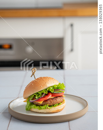 Meat burger with tomato, cheese, and lettuce on a plate on a table against a white kitchen Meat burger with tomato, cheese, and lettuce on a plate on a table against a white kitchen 138029365
